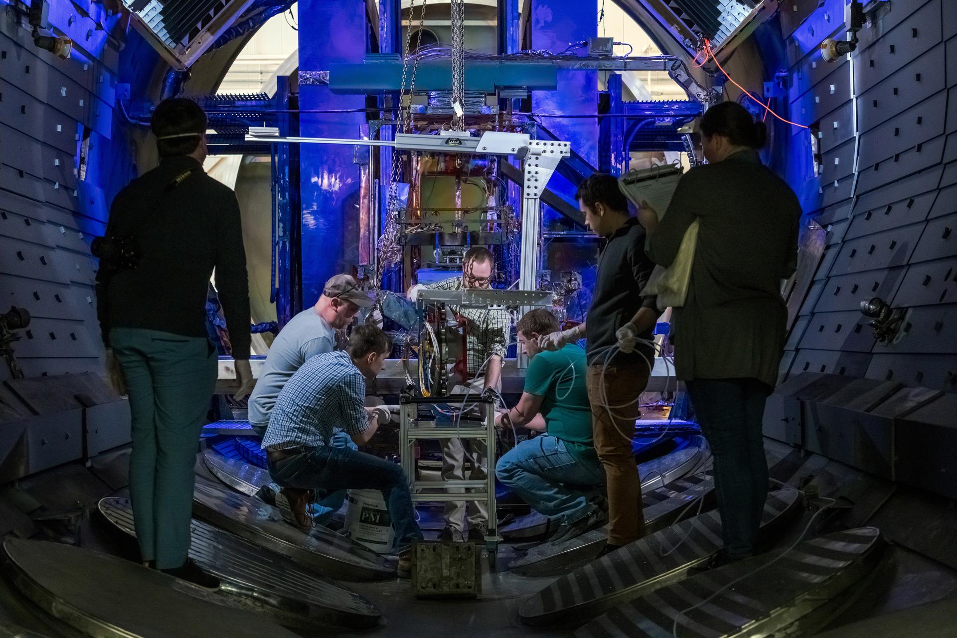 A group of people work together inside a vacuum chamber at NASA's Glenn Research Center to prepare an Advanced Electric Propulsion System (AEPS) thruster for testing. Most are kneeling or crouching around the thruster, which is mounted on a wheeled metal stand near the center of the chamber.