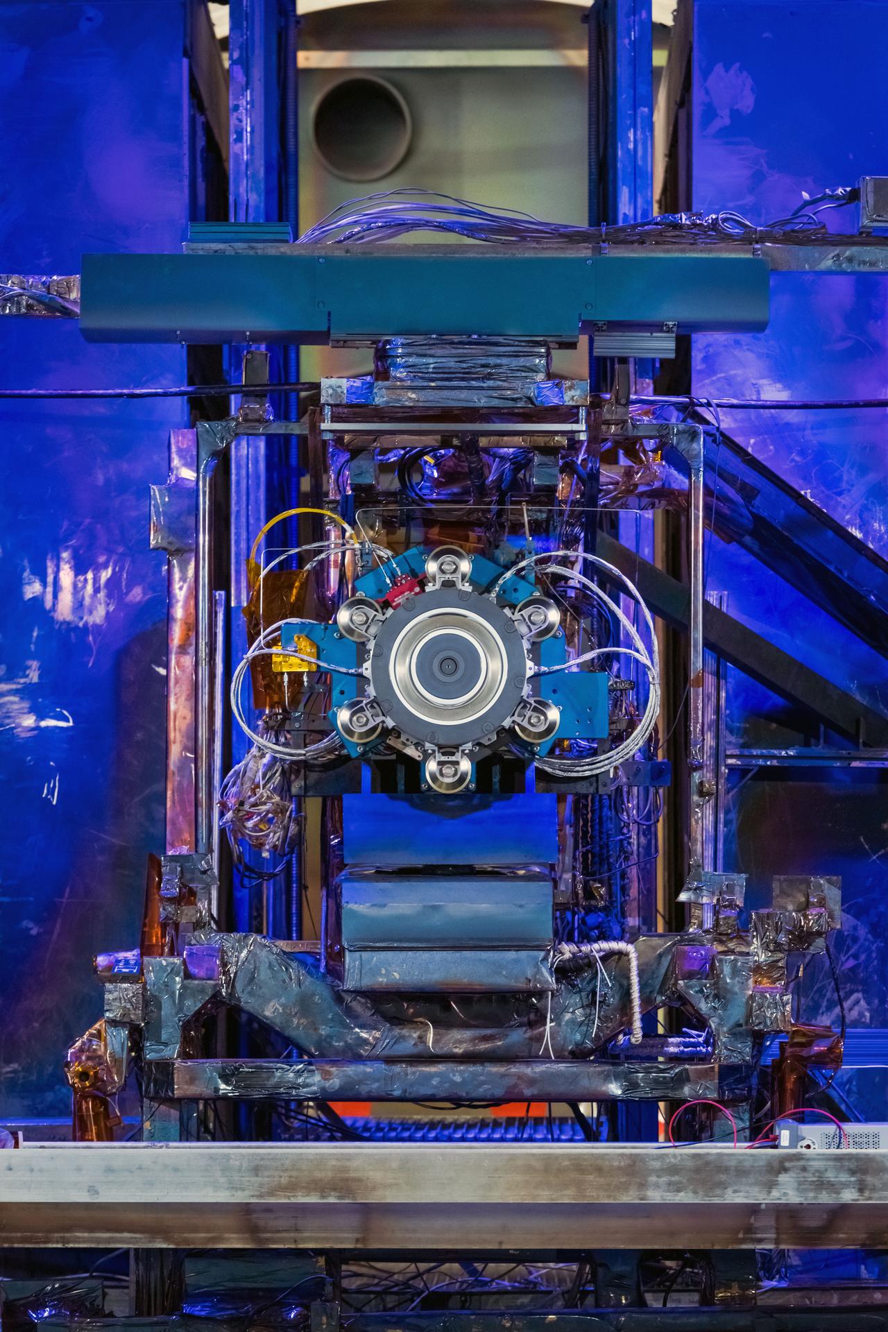 Close-up view of an Advanced Electric Propulsion System (AEPS) thruster mounted inside a vacuum chamber at NASA Glenn Research Center. The thruster is centered in the image, featuring a circular central component surrounded by six evenly spaced protrusions, with multiple cables and wires connected to its structure.