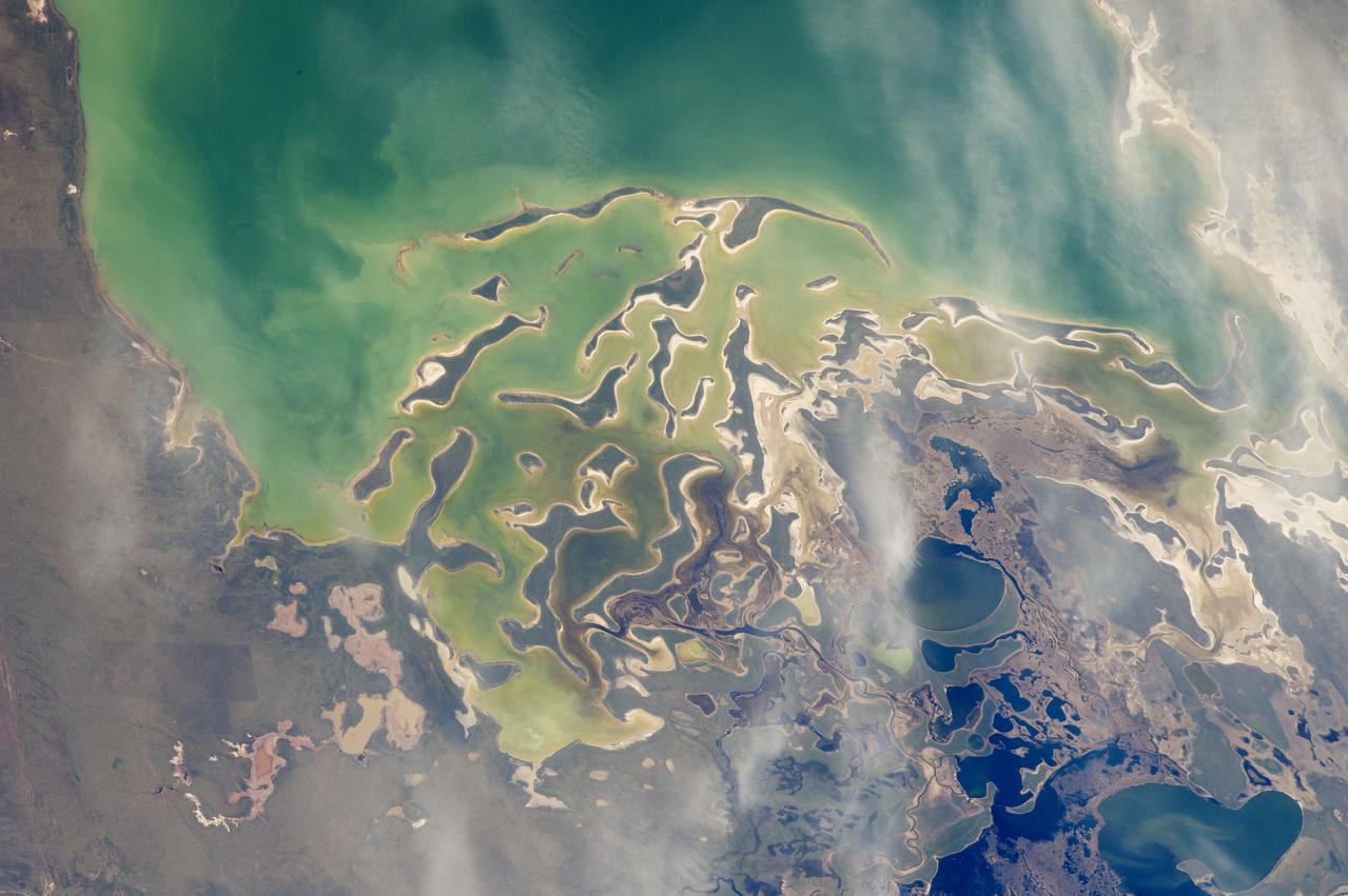 In orbit above the semi-desert grasslands in Kazakhstan, an astronaut aboard the International Space Station spotted one of the few features that stand out. Lake Tengiz is the only large lake (1590 square kilometers, 615 square miles) in northern Kazakhstan. Through white wisps of cloud, the crew member photographed the 50 kilometer-long eastern shore of the lake, with its thin, winding islands and white beaches. The islands and intervening waterways make a rich habitat for birds in this part of Asia. At least 318 species of birds have been identified at the lake; 22 of them are endangered. It is the northernmost habitat of the pink flamingo. The lake system is Kazakhstan’s first UNESCO World Heritage Site, and it has been declared a RAMSAR wetland site of international importance. Part of the richness of area is its complex hydrology. Fresh water enters the system via the Kulanutpes River, so there are small lakes (lower right) full of fresh water. But in this closed basin, the water in the main lake (top) slowly evaporates, becoming salty. Winds stir up bigger waves on the main lake, dispersing sediment and salt and making the water a cloudier and lighter blue-green. (Another astronaut photograph shows the entire lake system, while this story provides more information.) The strange shape of the islands is not easy to interpret. They may be drowned remnants of delta distributaries of the Kulanutpes River. Westerly winds probably have had a smoothing effect on the shorelines, especially in a shallow lake like Tengiz, which is only about 6 meters (20 feet) deep. The lake has an exciting history for people who follow space exploration. In 1976, a Soyuz spacecraft landed in the lake near the north shore (top right). The capsule crashed through the ice and sank during an October snowstorm when temperatures were -22°C (-8°F). Because of low power, the capsule was unheated and the crew was feared lost. It was many hours before the airtight capsule was located and divers could attach flotation tanks to get the capsule to the surface. It was then dragged ashore across the ice by helicopter. The rescue effort took nine hours before the crew was able to safely exit the capsule. Credit: <b><a href="http://www.earthobservatory.nasa.gov/" rel="nofollow"> NASA Earth Observatory</a></b> <b><a href="http://www.nasa.gov/audience/formedia/features/MP_Photo_Guidelines.html" rel="nofollow">NASA image use policy.</a></b> <b><a href="http://www.nasa.gov/centers/goddard/home/index.html" rel="nofollow">NASA Goddard Space Flight Center</a></b> enables NASA’s mission through four scientific endeavors: Earth Science, Heliophysics, Solar System Exploration, and Astrophysics. Goddard plays a leading role in NASA’s accomplishments by contributing compelling scientific knowledge to advance the Agency’s mission. <b>Follow us on <a href="http://twitter.com/NASAGoddardPix" rel="nofollow">Twitter</a></b> <b>Like us on <a href="http://www.facebook.com/pages/Greenbelt-MD/NASA-Goddard/395013845897?ref=tsd" rel="nofollow">Facebook</a></b> <b>Find us on <a href="http://instagrid.me/nasagoddard/?vm=grid" rel="nofollow">Instagram</a></b>