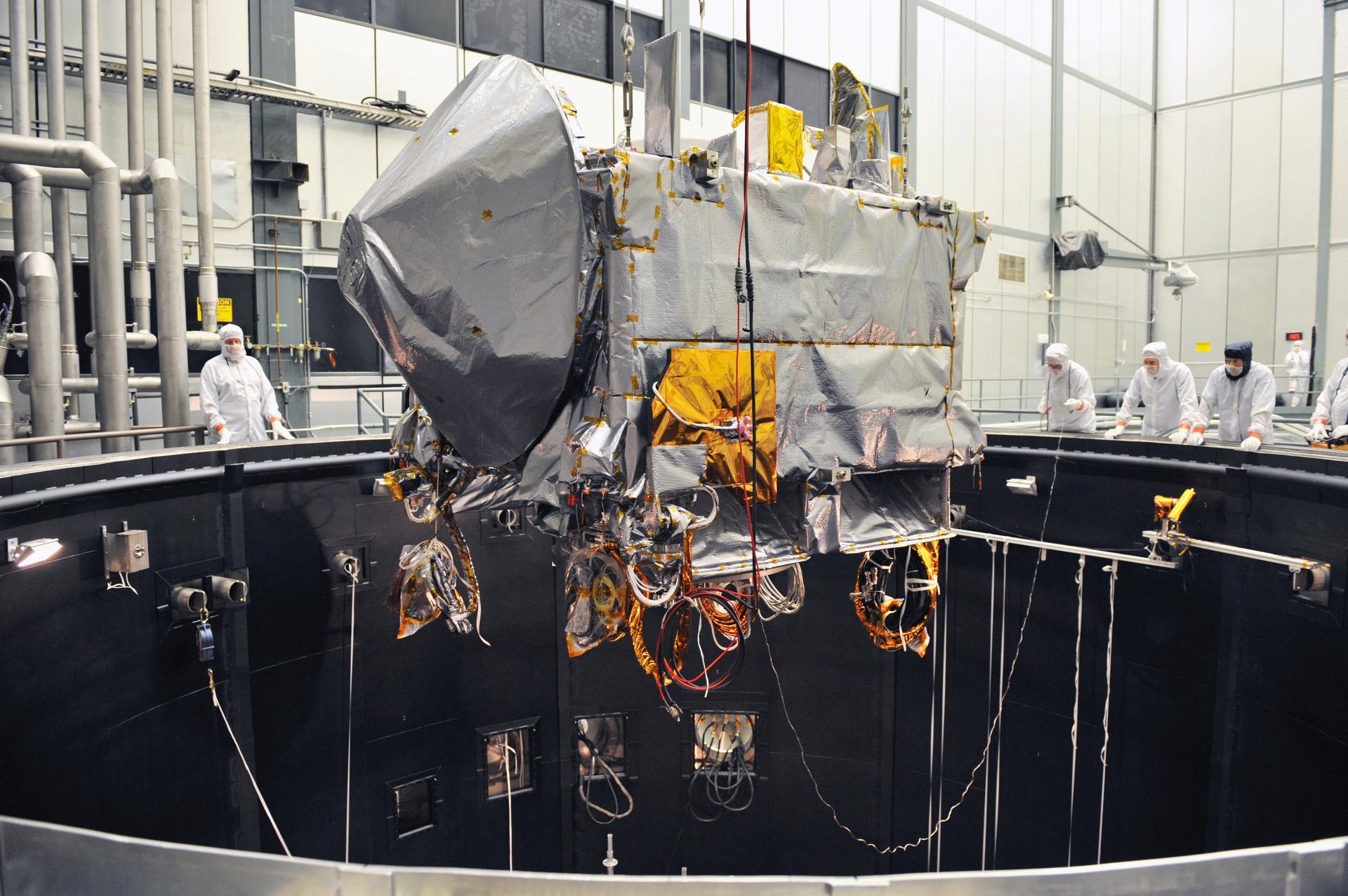 The OSIRIS-REx spacecraft is lowered into a black thermal vacuum chamber from above. The spacecraft is a rectangle shape, wrapped in silver blanketing. A squat cone on the front end is also wrapped in silver blanketing, placed like the head on a dog. Various wires extend from the spacecraft. People in white clean room suits watch from the edge of the chamber.