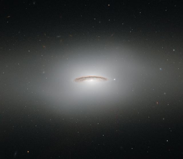 This neat little galaxy is known as NGC 4526. Its dark lanes of dust and bright diffuse glow make the galaxy appear to hang like a halo in the emptiness of space in this image from the NASA/ESA Hubble Space Telescope.  Although this image paints a picture of serenity, the galaxy is anything but. It is one of the brightest lenticular galaxies known, a category that lies somewhere between spirals and ellipticals. It has hosted two known supernova explosions, one in 1969 and another in 1994, and is known to have a colossal supermassive black hole at its center that has the mass of 450 million suns.  NGC 4526 is part of the Virgo cluster of galaxies. Ground-based observations of galaxies in this cluster have revealed that a quarter of these galaxies seem to have rapidly rotating disks of gas at their centers.  The most spectacular of these is this galaxy, NGC 4526, and its spinning disk of gas, dust, and stars reaches out uniquely far from its heart, spanning some seven percent of the galaxy's entire radius.  This disk is moving incredibly fast, spinning at more than 250 kilometers per second. The dynamics of this quickly whirling region were actually used to infer the mass of NGC 4526’s central black hole — a technique that had not been used before to constrain a galaxy’s central black hole.  This image was taken with Hubble's Wide Field and Planetary Camera 2 and the Advanced Camera for Surveys.  Credit: ESA/Hubble &amp; NASA, Acknowledgement: Judy Schmidt