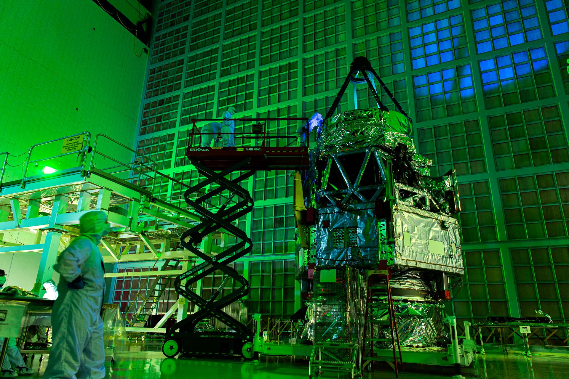 A low angle view of the Roman Space telescope. one person stands in bottom left of the frame watching as three others stand on a scissor lift with one shining a UV light onto the primary mirror as part of an inspection. The photograph is a long exposure which makes the entire image look green from ambient light and a large blue spot appears on the back filter wall due to the UV light reflecting off the primary mirror