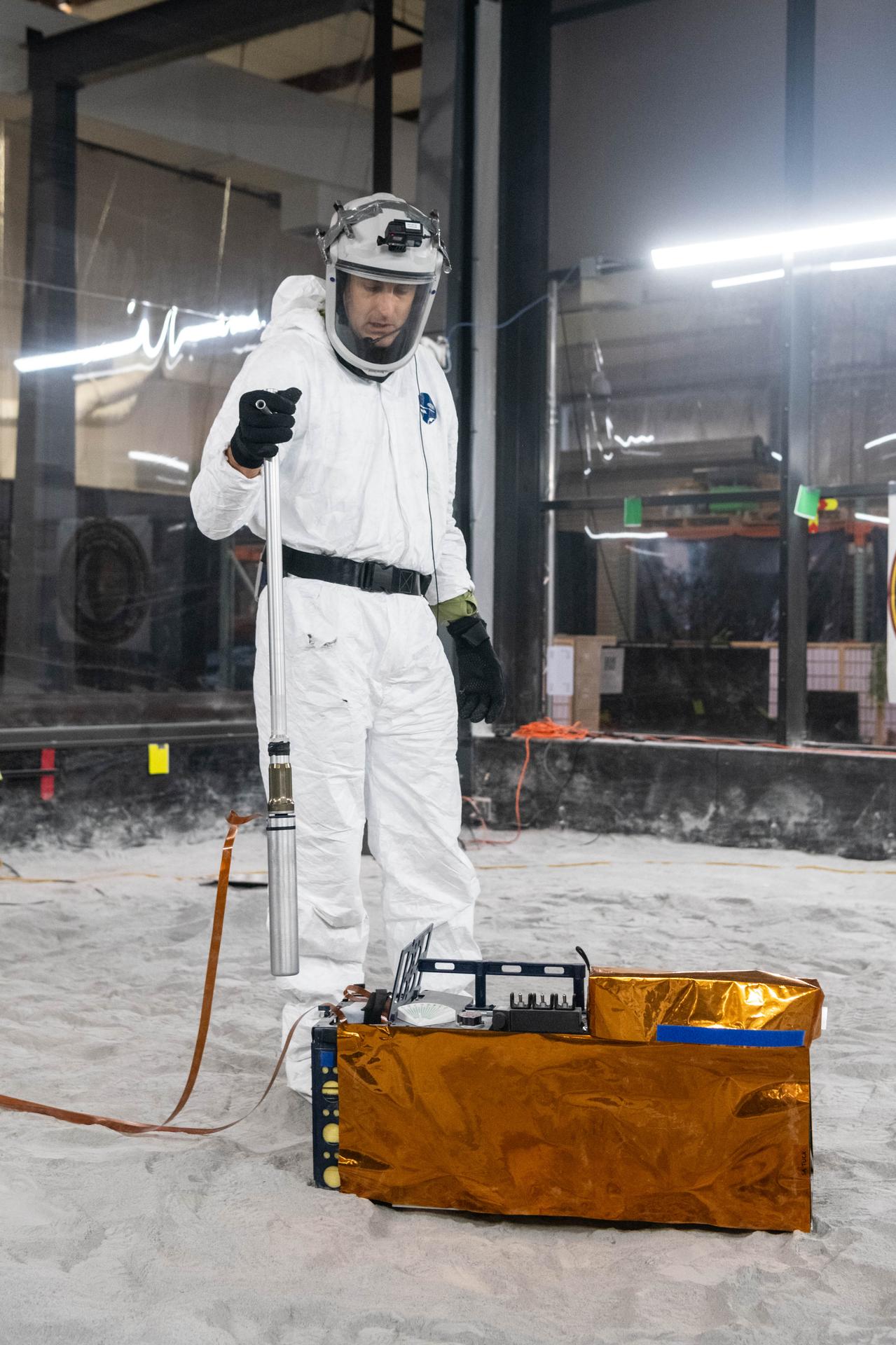 Artemis Scientist, Trevor Graff, performs instrument deployment exercises with a medium fidelity mock up of the Lunar Environment Monitoring Station (LEMS) in the lunar simulant bin at the Florida Space Institute Exolith Lab, Orlando, Fl., Aug 6, 2025. LEMS is a compact, autonomous, and self-sustaining seismometer suite designed to carry out continuous, long-term, monitoring of the lunar seismic environment at the South Polar region. Photo Credit: NASA/Katie Mellos.