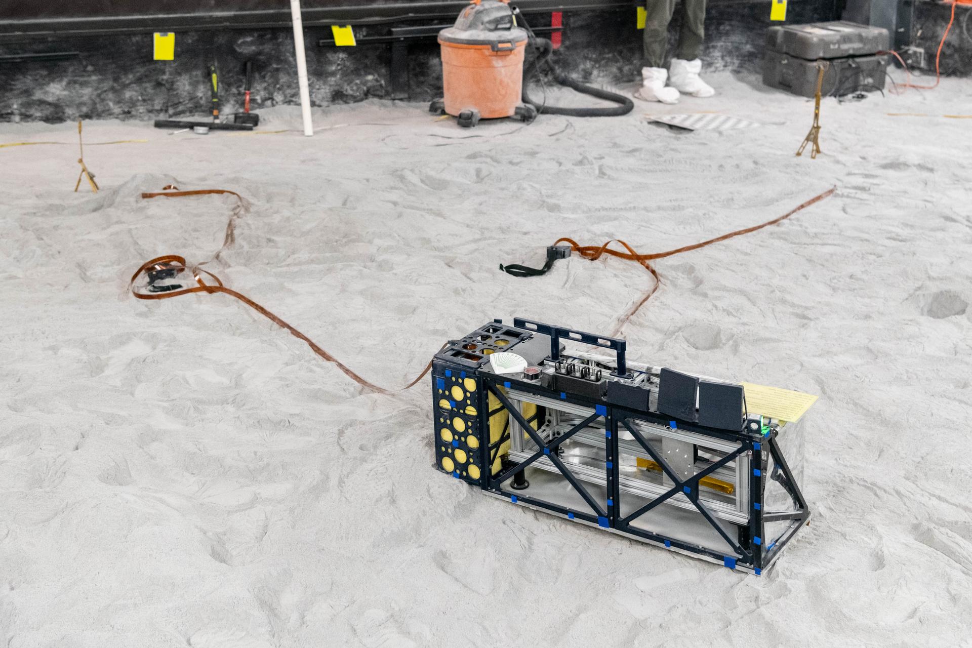 The Lunar Environment Monitoring Station (LEMS) medium fidelity instrument mock up is configured for instrument deployment exercises in the lunar simulant bin at the Florida Space Institute Exolith Lab, Orlando, Fl., Aug 6, 2025. LEMS is a compact, autonomous, and self-sustaining seismometer suite designed to carry out continuous, long-term, monitoring of the lunar seismic environment at the South Polar region. Photo Credit: NASA/Katie Mellos.