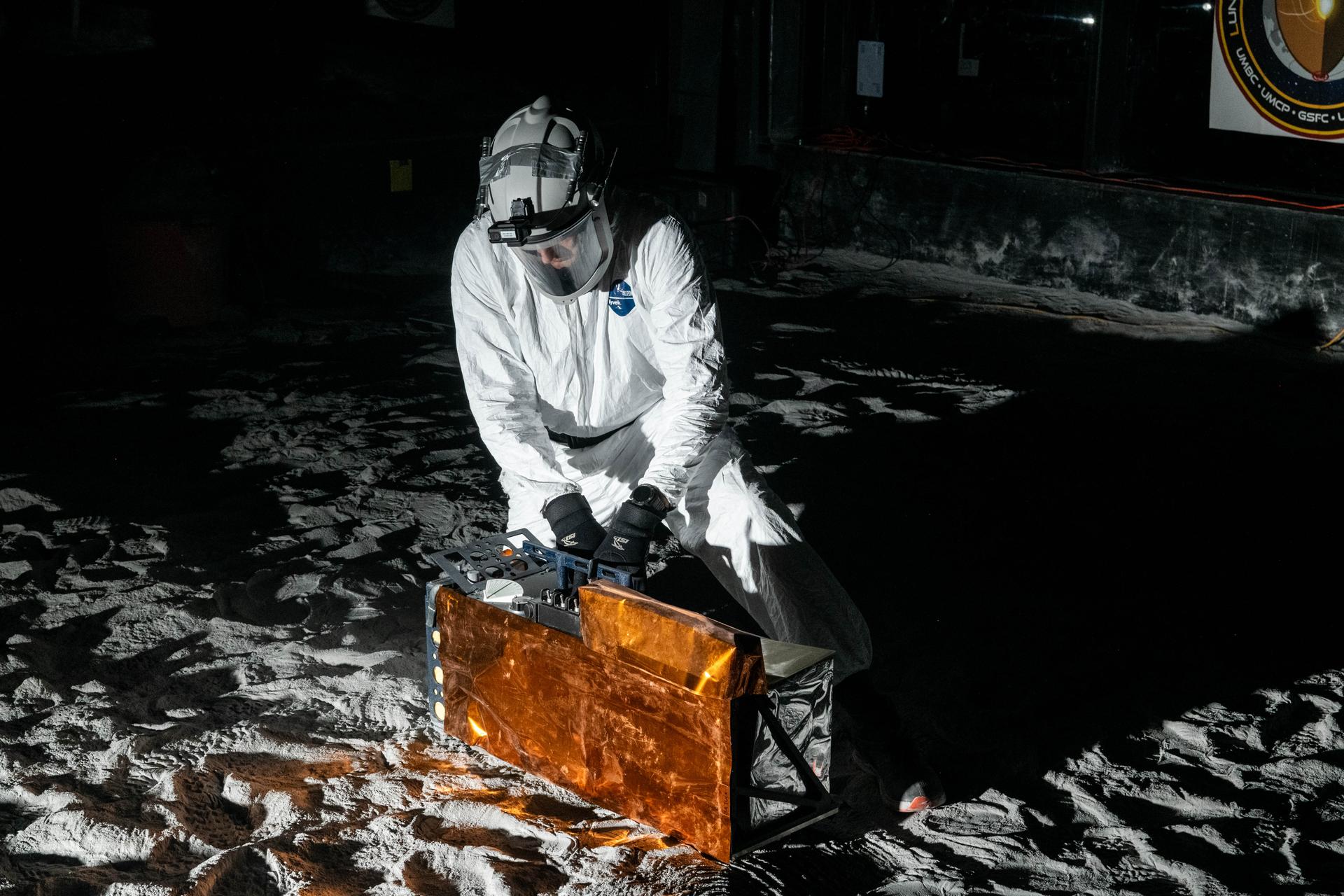 Artemis Scientist, Trevor Graff, performs instrument deployment exercises with a medium fidelity mock up of the Lunar Environment Monitoring Station (LEMS) in the lunar simulant bin at the Florida Space Institute Exolith Lab, Orlando, Fl., Aug 6, 2025. LEMS is a compact, autonomous, and self-sustaining seismometer suite designed to carry out continuous, long-term, monitoring of the lunar seismic environment at the South Polar region. Photo Credit: NASA/Katie Mellos.
