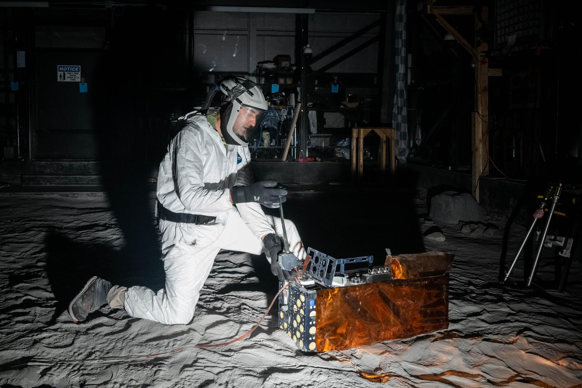 Artemis Scientist, Trevor Graff, performs instrument deployment exercises with a medium fidelity mock up of the Lunar Environment Monitoring Station (LEMS) in the lunar simulant bin at the Florida Space Institute Exolith Lab,  Orlando, Fl., Aug 6, 2025. LEMS is a compact, autonomous, and self-sustaining seismometer suite designed to carry out continuous, long-term, monitoring of the lunar seismic environment at the South Polar region. Photo Credit: NASA/Katie Mellos.