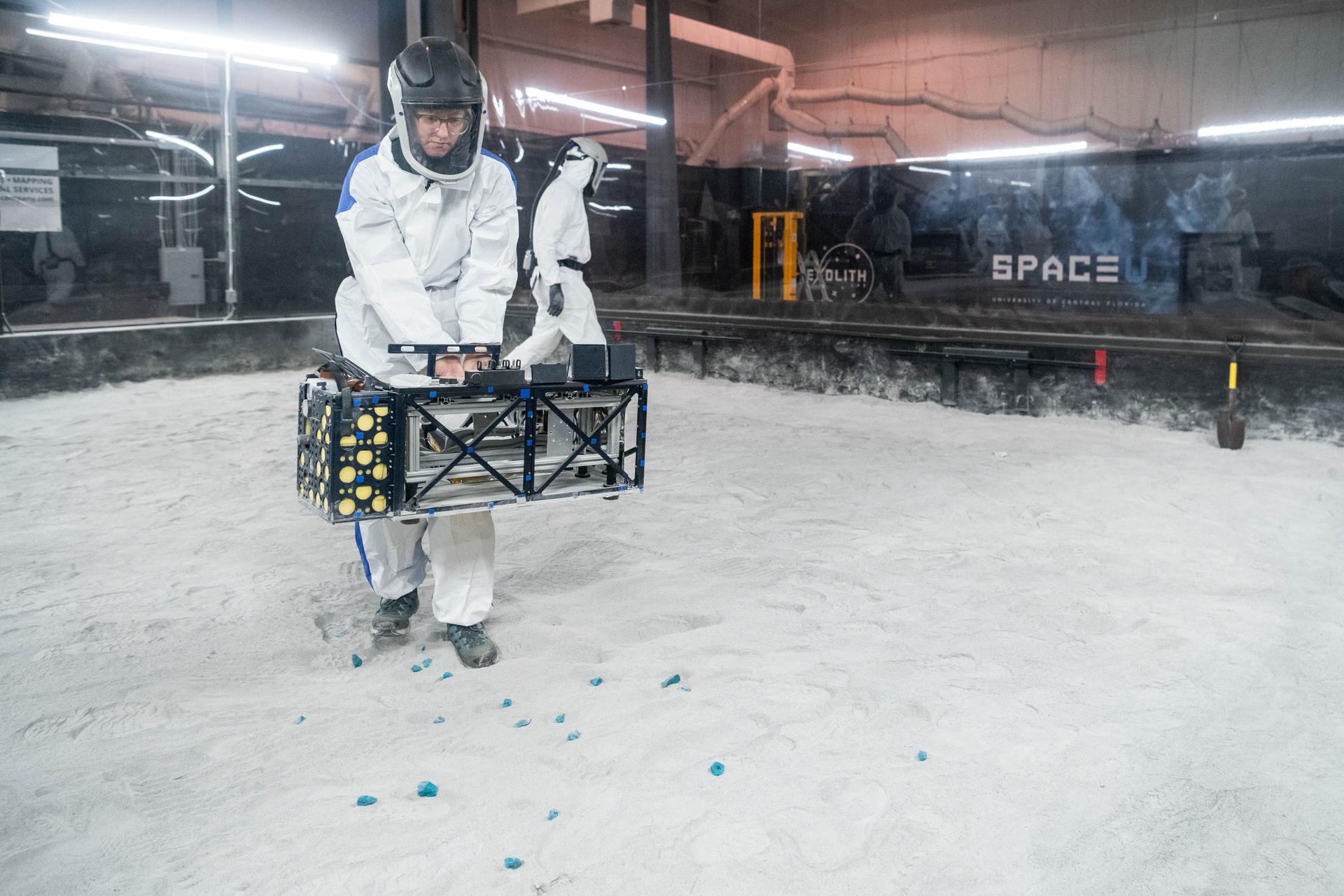 Naoma McCall, Science Co-Investigator, Deployment Lead, performs instrument deployment exercises with a medium fidelity mock up of the Lunar Environment Monitoring Station (LEMS) in the lunar simulant bin at the Florida Space Institute Exolith Lab, Orlando, Fl., Aug 7th, 2025. LEMS is a compact, autonomous, and self-sustaining seismometer suite designed to carry out continuous, long-term, monitoring of the lunar seismic environment at the South Polar region. Photo Credit: NASA/Katie Mellos.