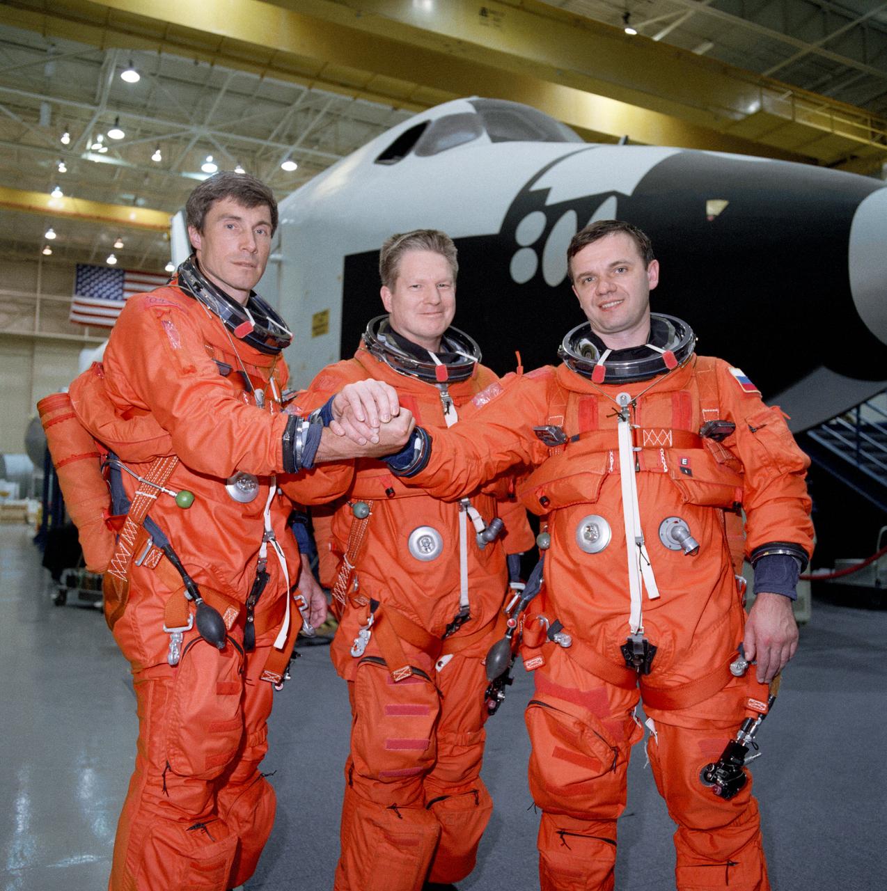 Photographic documentation showing the first crew of the ISS posing together in bldg. 9N during descent training, wearing orange Launch & Entry Suits (LES) and clasping right hands in a sign of unity. From left to right: Sergei Krikalev, William Shepherd and Yuri Gidzenko.