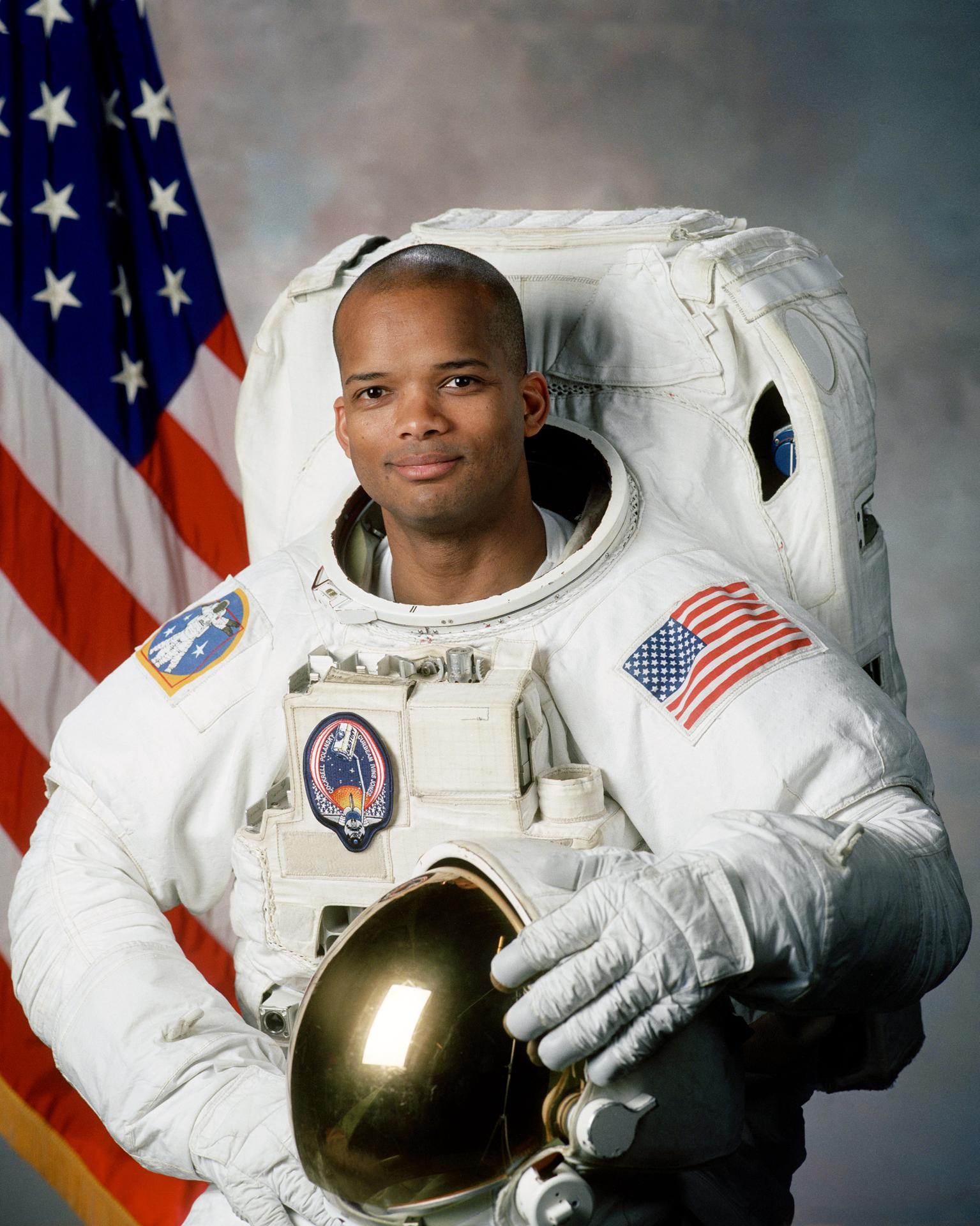 A man smiles, wearing a spacesuit, and stands in front of the American flag. He has his helmet off and sitting before him.