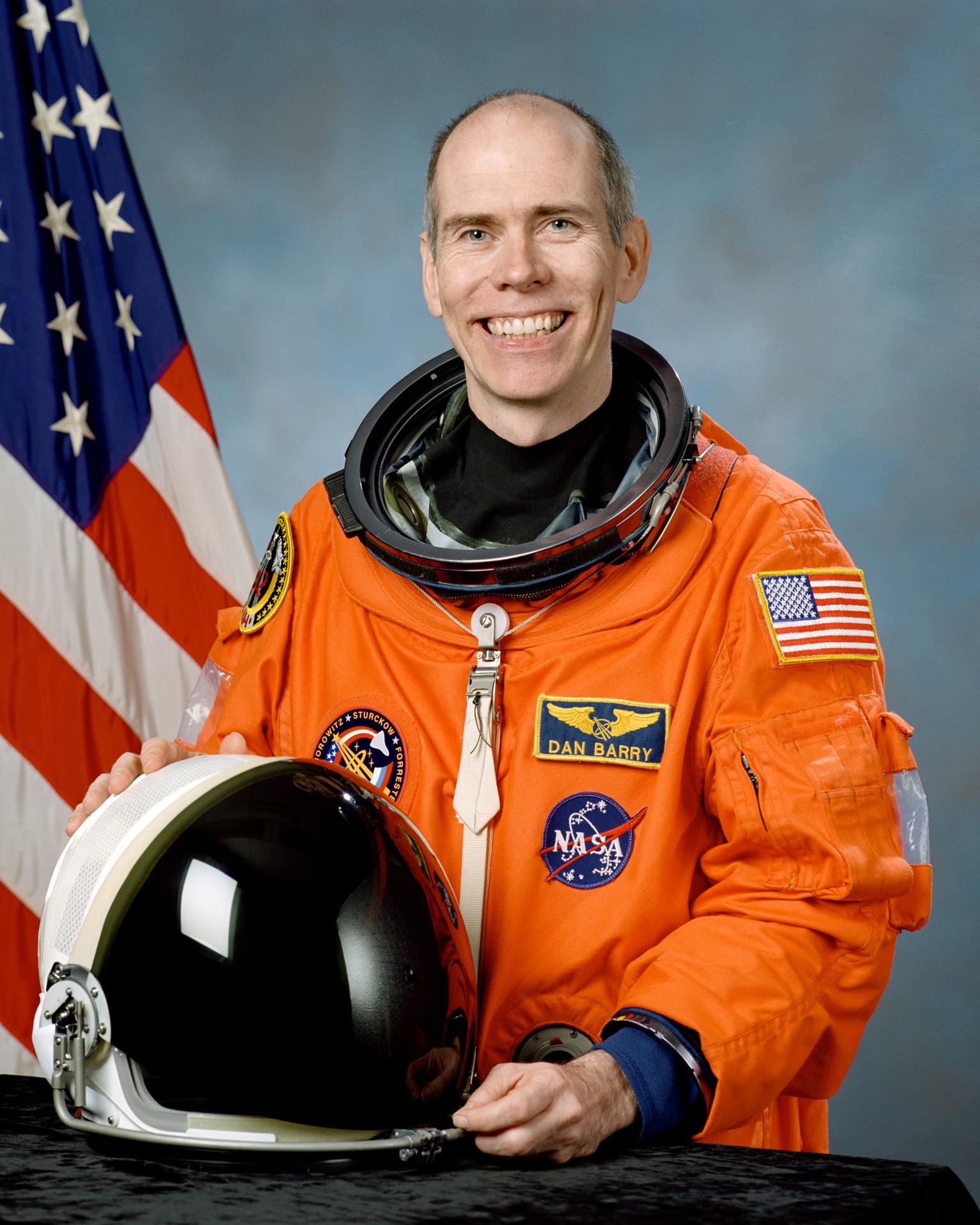 A man sits before the American flag in an astronaut suite, with his helmet off