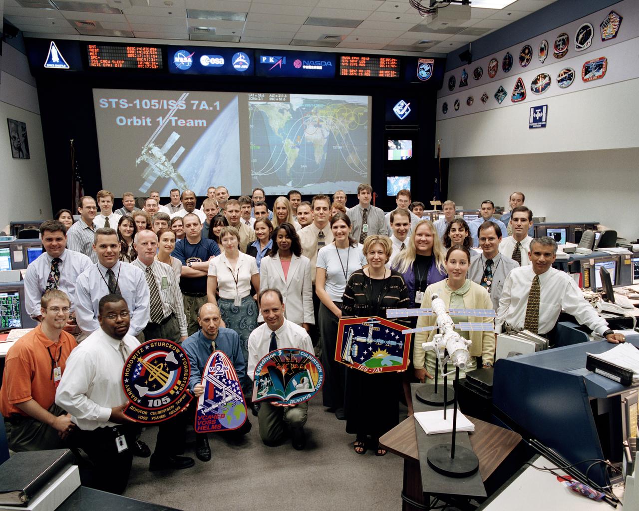 JSC2001-02229 (16 August 2001) --- The members of the STS-105/ISS 7A.1 Orbit 1 team pose for a group portrait in the International Space Station (ISS) flight control room (BFCR) in Houston&#0146;s Mission Control Center (MCC).  Flight director Mark Ferring is kneeling as he holds the Expedition Three mission logo.  Astronaut Stephanie D. Wilson, ISS spacecraft communicator (CAPCOM), is standing behind Ferring.