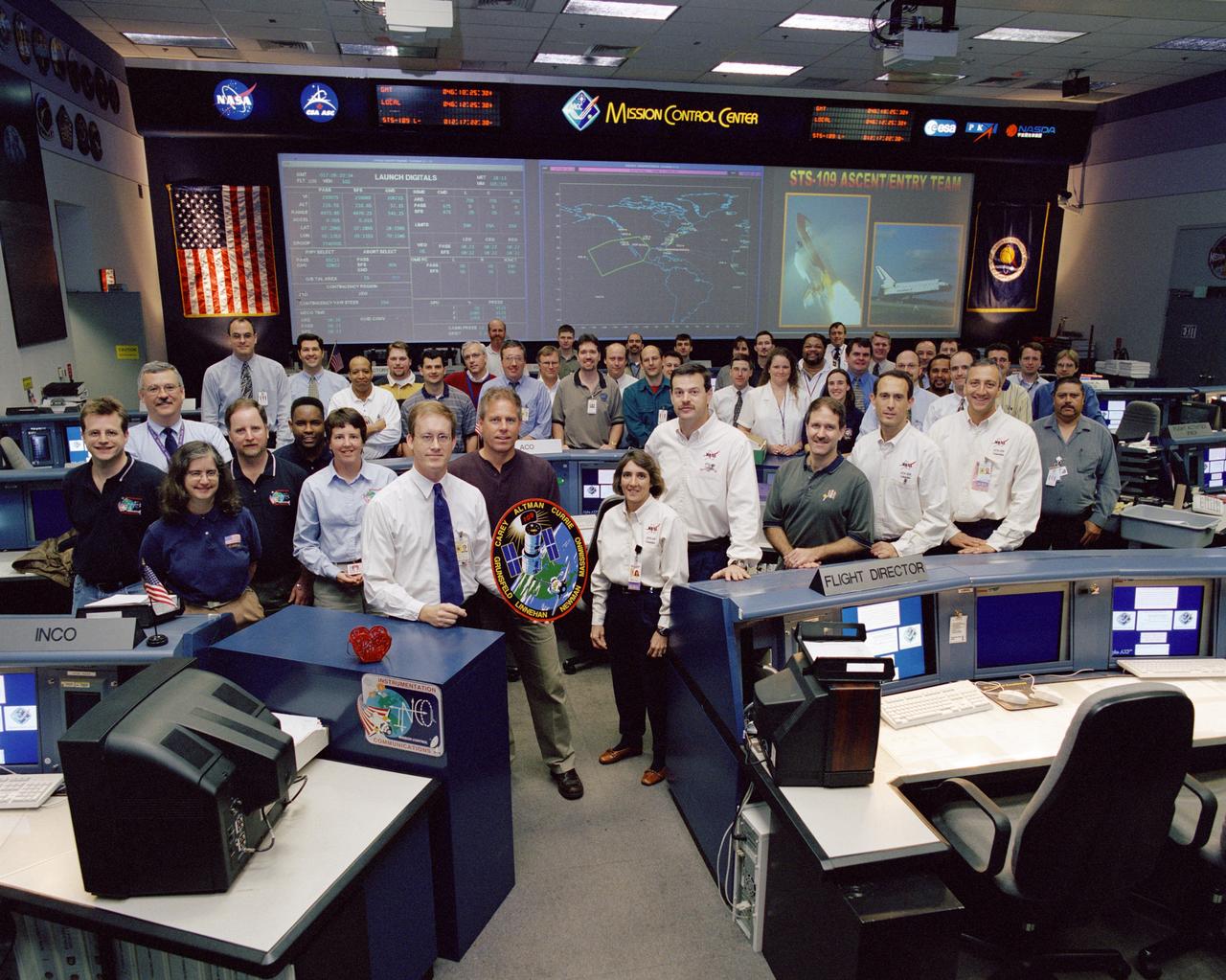 JSC2002-00514 (February 2002)--- The STS-109 flight crew poses with the ascent and entry shift team in the Shuttle Flight Control Room of the Johnson Space Center's Mission Control Center.  Flight Director John Shannon holds the mission insignia.  Members of the flight crew are astronauts Scott D. Altman, commander; Duane G. Carey, pilot; John M. Grunsfeld, payload commander; and James H. Newman, Nancy J. Currie, Richard M. Linnehan and Michael J. Massimino, all mission specialists.  Currie stands to the right of the logo, followed  by, left to right,  Altman, Grunsfeld, Newman and Massimino.  Linnehan and Carey are not pictured.