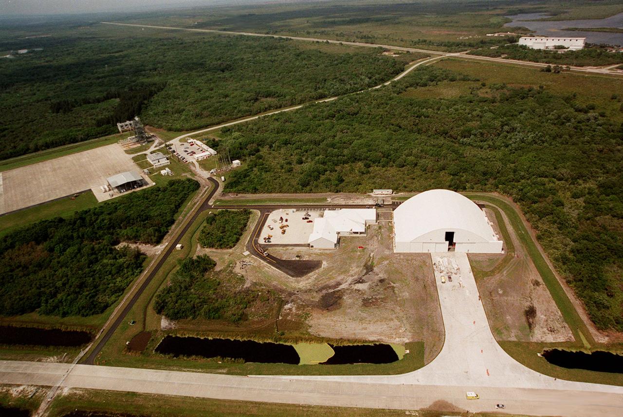 Looking east in this aerial photo can be seen several facilities at KSC. On the left is the parking tarmac next to the runway at the Shuttle Landing Facility. Astronaut Rd. runs from it toward the upper right corner, connecting with Kennedy Parkway North. The white building, upper right) is the Apollo/Saturn V Center. At the center of the photo is the remote launch vehicle (RLV) hangar, still under construction, at the south end of the Shuttle Landing Facility. Next to the multi-purpose RLV hangar are facilities for related ground support equipment and administrative/technical support. At the bottom of the photo is the tow-way road which connects the runway with the Orbiter Processing Facility.