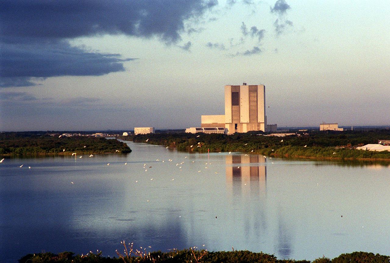 In the early morning, flocks of birds soar over the Banana River, whose waters reflect the Vehicle Assembly Building , bathed in a pink glow from post-dawn light. Next to the VAB, on the left, is the Launch Control Center. The rectangular building closer to the water at left is the Operations Support Building. At right is the Rotation/Processing Facility. The birds are a common sight at KSC since the Center shares a boundary with the Merritt Island National Wildlife Refuge. The Refuge encompasses 92,000 acres that are a habitat for more than 331 species of birds, 31 mammals, 117 fishes, and 65 amphibians and reptiles. The marshes and open water of the refuge provide wintering areas for 23 species of migratory waterfowl, as well as a year-round home for great blue herons, great egrets, wood storks, cormorants, brown pelicans and other species of marsh and shore birds, as well as a variety of insects.