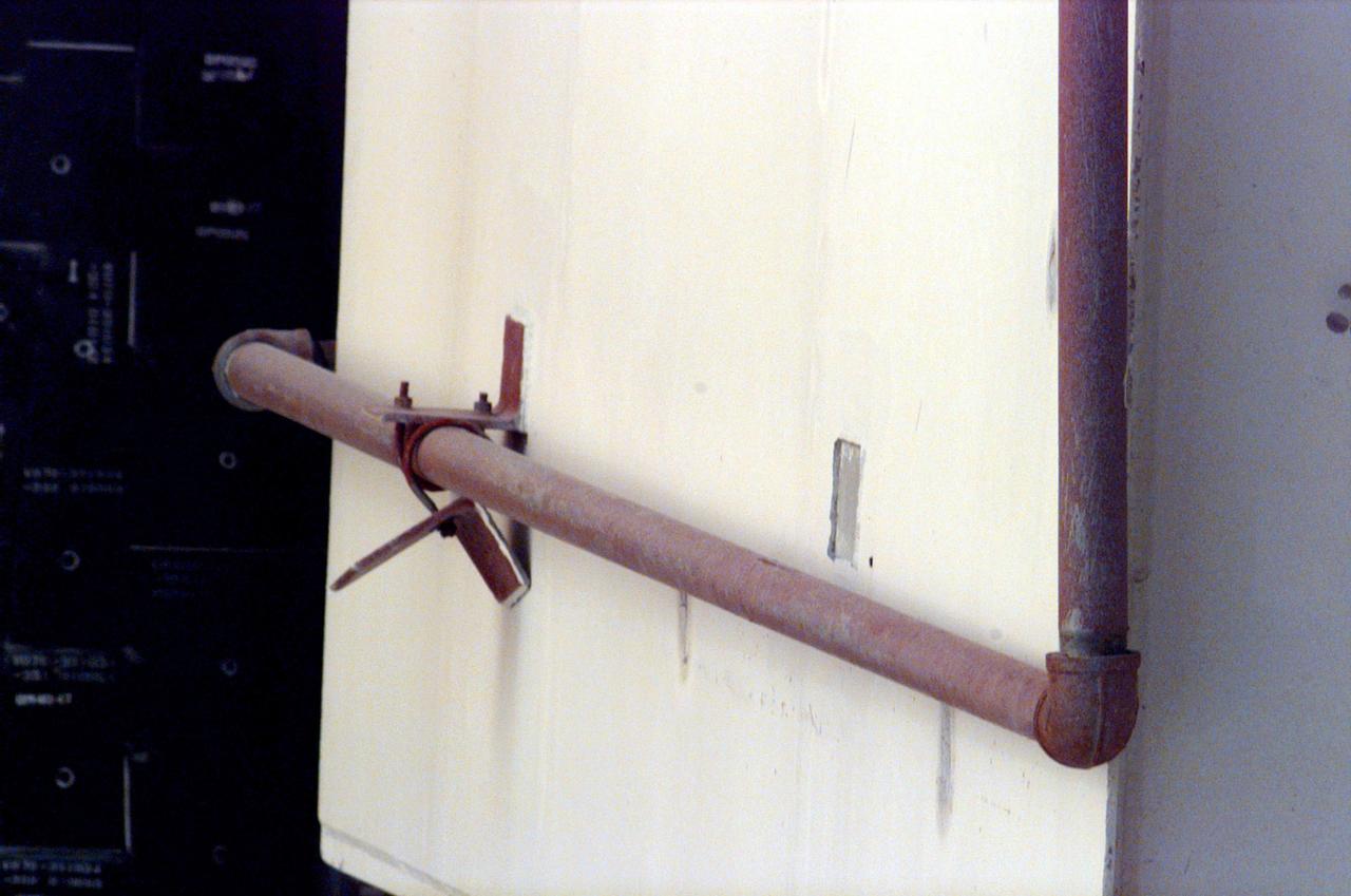 This loose bracket, observed hanging down from the side of the White Room at Launch Pad 39B, delayed loading of Endeavour's external tank by several hours to allow technicians to remove it. A "U" bolt connects the bracket to a fire suppression water line attached to the exterior of the White Room. The loose bolt could have possibly created a debris hazard