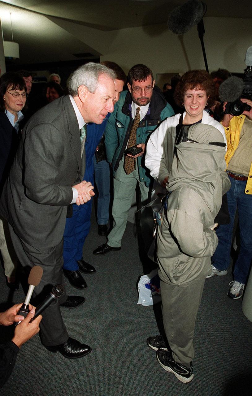 NASA Administrator Dan Goldin (left) listens intently to 10-year-old Jonathan Pierce (right), who is garbed in a protective cooling suit designed by NASA. Behind Goldin is astronaut Doug Wheelock; behind Jonathan is his mother, Penny. Jonathan suffers from erythropoietic protoporphyria, a rare condition that makes his body unable to withstand ultraviolet rays. The suit allows him to be outside during the day, which would otherwise be impossible. Jonathan's trip was funded by the Make-A-Wish Foundation and included a visit to Disney World. He and his family were among a dozen VIPs at KSC to view the launch of STS-99