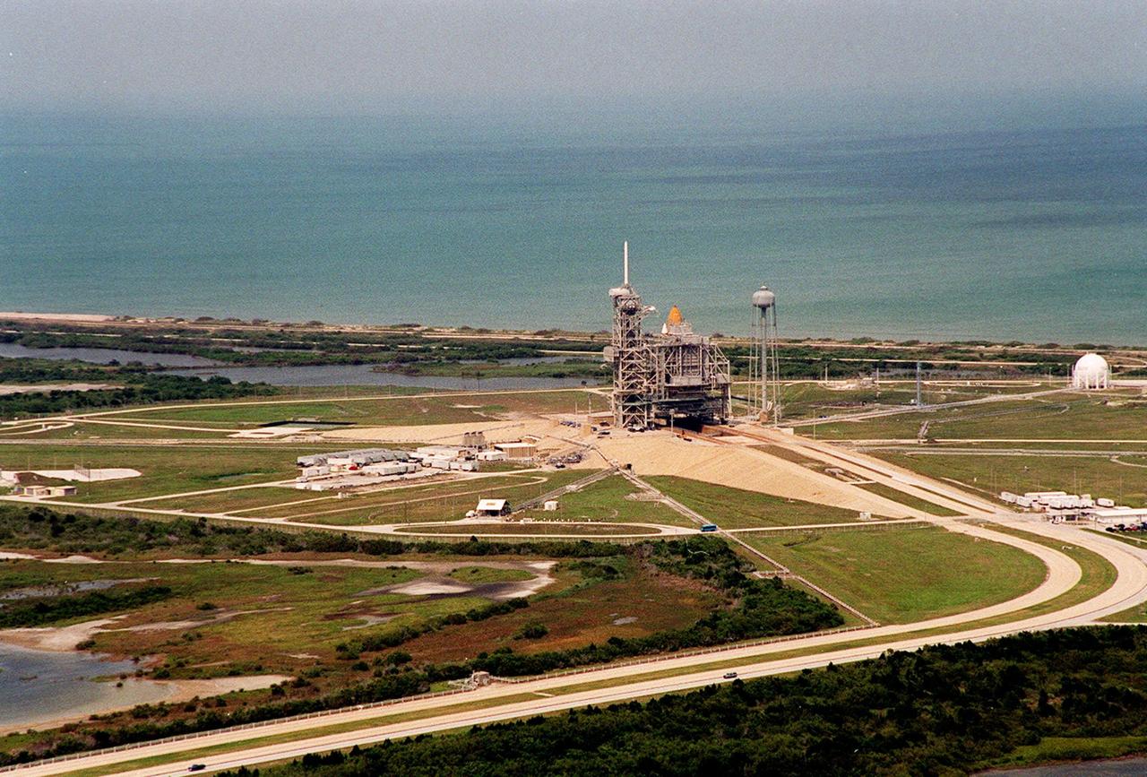 This aerial view captures KSC's Launch Pad 39A, with the rotating service structure closed around Shuttle Atlantis. Just visible in the center is the top of the orange-colored external tank. On the left side towers the white 80-foot fiberglass lightning mast; on the right is the 300,000-gallon water tower used for sound suppression during launch. The launch pad abuts the Atlantic Ocean on the east, seen here in the background