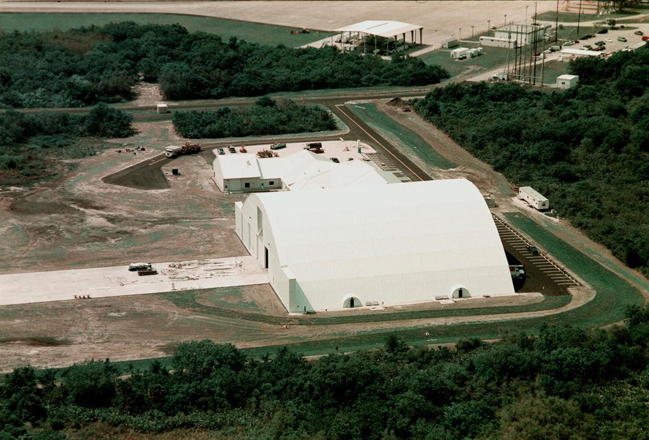 This aerial photo focuses on the remote launch vehicle (RLV) hangar, still under construction. It sits at the south end of the Shuttle Landing Facility. Adjacent to the multi-purpose RLV hangar (above it) are facilities for related ground support equipment and administrative/technical support. The top of the photo captures a portion of the parking tarmac near the runway