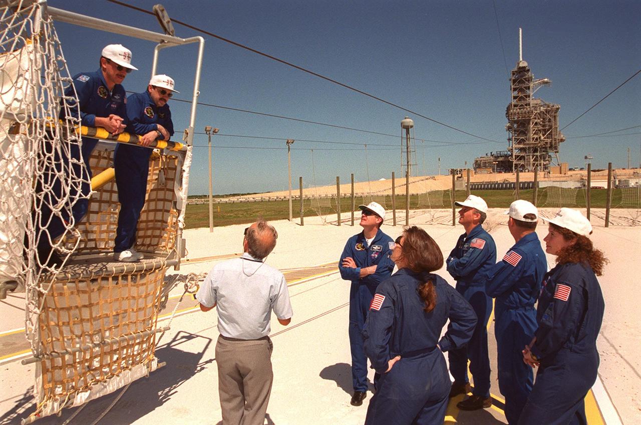 KENNEDY SPACE CENTER, Fla. -- During emergency egress training at Launch pad 39A, STS-101 Pilot Scott J. "Doc" Horowitz and Mission Specialist Yury Usachev stand in the slidewire basket at the landing zone. The Safety Egress officer (left on ground) provides training on use of the basket in the event the crew needed to exit quickly from the fixed service structure (background) at the 195-foot level. The rest of the crew, clockwise at right, are Commander James D. Halsell Jr. and Mission Specialists Jeffrey N. Williams, James Voss, Susan Helms, and (back to the camera) Mary Ellen Weber. The training is part of Terminal Countdown Demonstration Test (TCDT) activities that include a simulated launch countdown and familiarization with the payload. During their mission to the International Space Station, the STS-101 crew will be delivering logistics and supplies, plus preparing the Station for the arrival of the Zvezda Service Module, expected to be launched by Russia in July 2000. Also, the crew will conduct one space walk to perform maintenance on the Space Station. This will be the third assembly flight for the Space Station. STS-101 is scheduled to launch April 24 at 4:15 p.m. from Launch Pad 39A