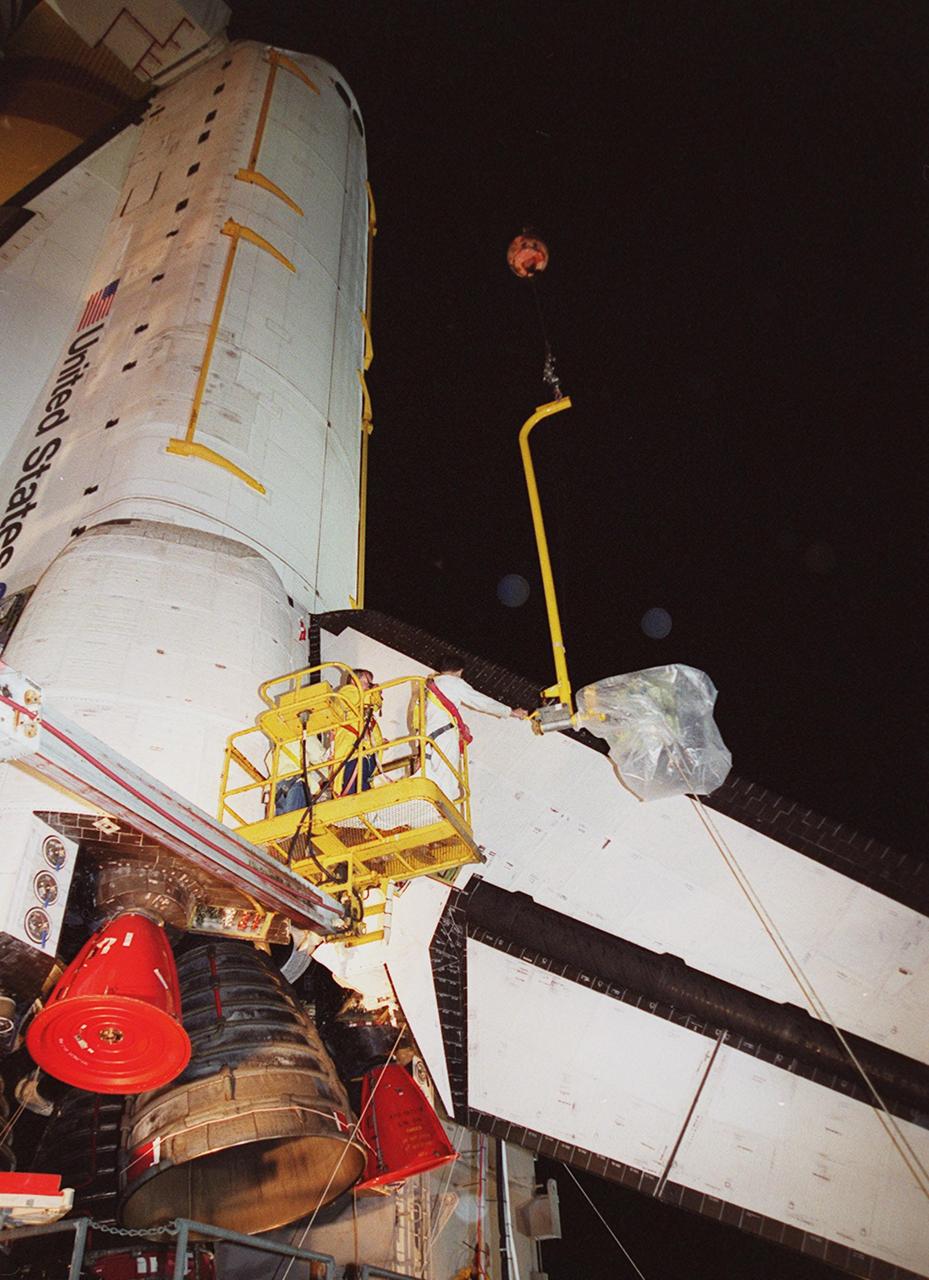 KENNEDY SPACE CENTER, FLA. -- At Launch Pad 39A, a worker reaches toward the plastic-covered replacement Power Drive Unit (PDU) for Space Shuttle Atlantis as it is lifted by crane toward the tail. The PDU controls the rudder/speed brake on the orbiter. Atlantis is scheduled to lift off April 24 at 4:15 p.m. EDT on mission STS-101, the third flight to the International Space Station. The primary mission is to carry logistics and supplies to the Space Station, plus the crew will be preparing the Station for the arrival of the Zvezda Service Module, expected to be launched by Russia in July 2000