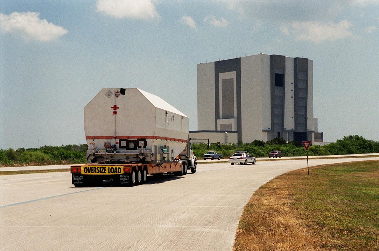 After its arrival at the Shuttle Landing Facility, the crated Tracking and Data Relay Satellite (TDRS-H) is transported past the Vehicle Assembly Building (in the background) to the Spacecraft Assembly and Encapsulation Facility (SAEF-2) for testing. The TDRS is one of three (labeled H, I and J) being built in the Hughes Space and Communications Company Integrated Satellite Factory in El Segundo, Calif. The latest TDRS uses an innovative springback antenna design. A pair of 15-foot-diameter, flexible mesh antenna reflectors fold up for launch, then spring back into their original cupped circular shape on orbit. The new satellites will augment the TDRS system’s existing Sand Ku-band frequencies by adding Ka-band capability. TDRS will serve as the sole means of continuous, high-data-rate communication with the space shuttle, with the International Space Station upon its completion, and with dozens of unmanned scientific satellites in low earth orbit. The TDRS is scheduled to be launched from CCAFS June 29 aboard an Atlas IIA/Centaur rocket