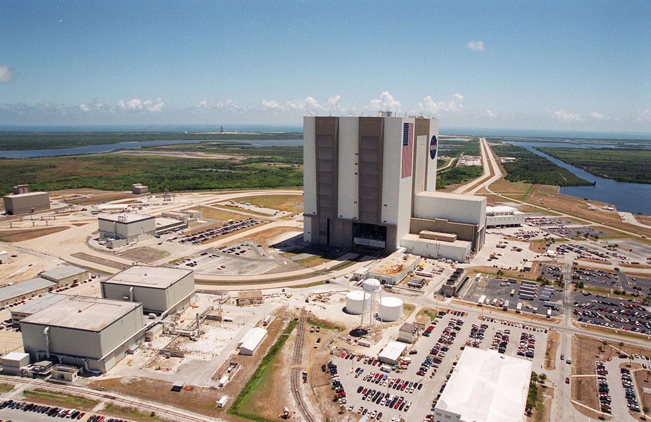 This aerial photo captures many of the facilities involved in Space Shuttle launches. At center is the Vehicle Assembly Building (VAB). The curved road on the near side is the newly restored crawlerway leading into the VAB high bay 2, where a mobile launcher platform/crawler-transporter currently sits. The road restoration and high bay 2 are part of KSC’s Safe Haven project, enabling the storage of orbiters during severe weather. The road circles around the Orbiter Processing Facility 3 (OPF-3) at left center. OPF1 and OPF-2 are just below the curving road. The crawlerway also extends from the east side of the VAB out to the two launch pads, only one visible to the left of the VAB. In the distance is the Atlantic Ocean. To the right of the far crawlerway is the turn basin, into which ships tow the barge for offloading new external tanks from Louisiana