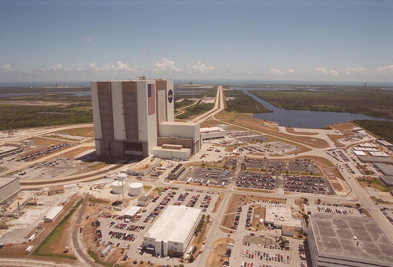 This aerial photo captures many of the facilities involved in Space Shuttle launches. At center is the Vehicle Assembly Building (VAB). The curved road on the near side is the newly restored crawlerway leading into the VAB high bay 2, where a mobile launcher platform/crawler-transporter currently sits. The road restoration and high bay 2 are part of KSC’s Safe Haven project, enabling the storage of orbiters during severe weather. The crawlerway also extends from the east side of the VAB out to the two Space Shuttle launch pads. In the distance is the Atlantic Ocean. To the right of the far crawlerway is the turn basin, into which ships tow the barge for offloading new external tanks from Louisiana