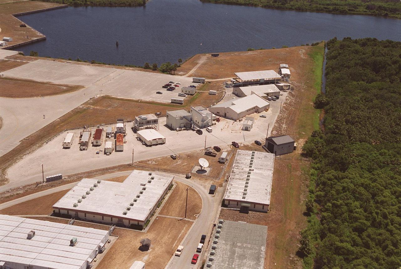 This aerial view is of the buildings that comprise the NASA Press Site in the Launch Complex 39 area. The first large building on the right (closest to the water) is the grandstand from which media representatives view the Space Shuttle launches. The building next to it houses the auditorium from which NASA press briefings are broadcast. To its immediate left is the NASA News Center which includes the offices of the NASA spokesmen at Kennedy Space Center. The buildings and trailers to the left of the News Center are assigned to various television stations and news services
