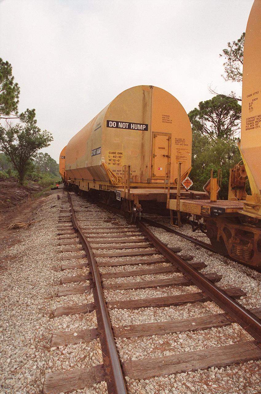 After being involved in a minor derailment incident during a routine movement on the tracks, rail cars carrying solid rocket booster segments sit idle. The rail cars were being moved as part of a standard operation to “order” the cars, placing them into a proper sequence for upcoming segment processing activities. The rear wheels of one car and the front wheels of the car behind it slid off the tracks while passing through a railway switch onto a siding. They were traveling approximately 3 miles per hour at the time, about normal walking speed. No damage occurred to the SRB segments, or to the devices that secure the segments to the rail cars. The incident occurred on KSC property, just north of the NASA Causeway in the KSC Industrial Area