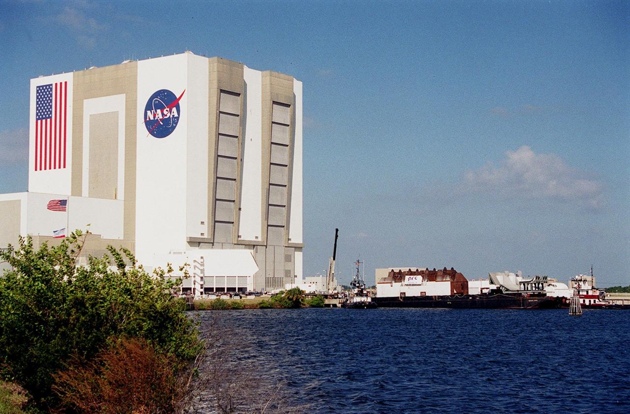 In the turn basin near the Vehicle Assembly Building (left) a launch table is docked, waiting for transfer to Launch Complex 37B, Cape Canaveral Air Force Station. The table was fabricated by Jered Industries in Georgia in support of the Delta Evolved Expendable Launch Vehicle (EELV) program, known as Delta IV. It was floated on a barge down the Intercoastal Waterway, through the Barge Canal to the turn basin. The table is approximately 70 feet long, 40 feet wide and 50 feet high, and weighs about 600,000 pounds. Accompanying the launch table on the barge are flame deflectors, which are also to be erected on pad 37B