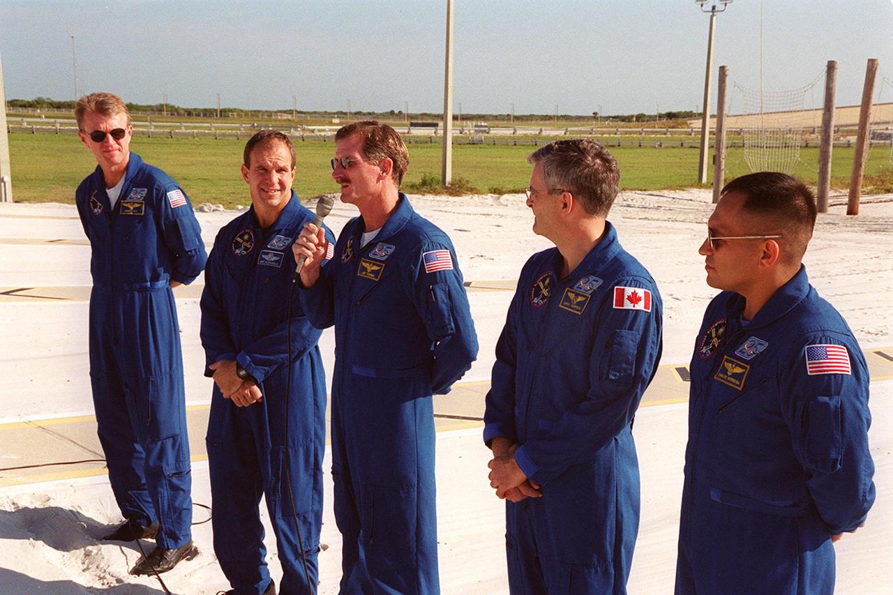 KENNEDY SPACE CENTER, Fla. -- From the slidewire landing zone at Launch Pad 39B, STS-97 Mission Specialist Joe Tanner (center, with microphone) speaks to the press about his extravehicular activity (EVA) during the mission. With him are the rest of the crew, Commander Brent Jett and Pilot Mike Bloomfield on the left and Mission Specialists Marc Garneau and Carlos Noriega on the right. The crew is at KSC to take part in Terminal Countdown Demonstration Test activities that include emergency egress training, familiarization with the payload, and a simulated launch countdown. Visible in the background are the solid rocket booster and external tank on Space Shuttle Endeavour. Mission STS-97is the sixth construction flight to the International Space Station. Its payload includes the P6 Integrated Truss Structure and a photovoltaic (PV) module, with giant solar arrays that will provide power to the Station. The mission includes two spacewalks to complete the solar array connections. STS-97 is scheduled to launch Nov. 30 at about 10:05 p.m. EST