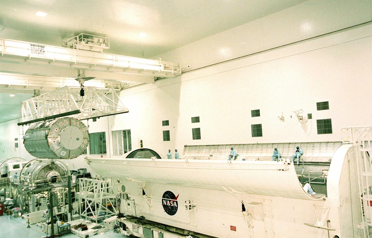 Workers wait inside the payload canister (right) for the U.S. Lab Destiny (left) to finish its move to the canister via overhead crane. Once installed inside the canister, Destiny will be transferred to Launch Pad 39A and lifted to the payload changeout room. From there it will be moved into the payload bay of the orbiter Atlantis. Destiny is the primary payload on mission STS-98, scheduled for launch no earlier than Jan. 19, 2001