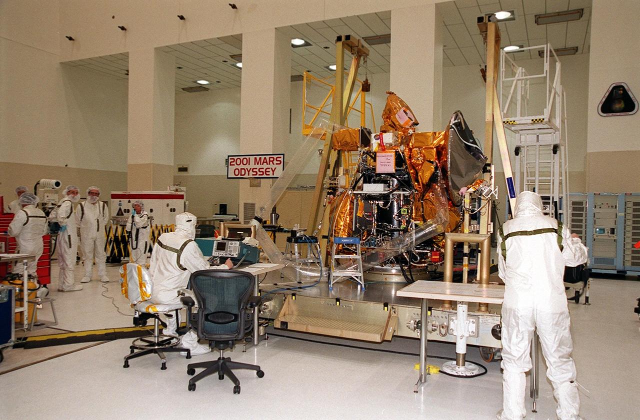 The 2001 Mars Odyssey Orbiter sits in the Spacecraft Assembly and Encapsulation Facility (SAEF 2) while components undergo testing. Workers in the foreground check instruments during testing of the UHF antenna. Scheduled for launch April 7, 2001, the orbiter contains three science instruments: THEMIS, the Gamma Ray Spectrometer (GRS), and the Mars Radiation Environment Experiment (MARIE). THEMIS will map the mineralogy and morphology of the Martian surface using a high-resolution camera and a thermal infrared imaging spectrometer. The GRS will achieve global mapping of the elemental composition of the surface and determine the abundance of hydrogen in the shallow subsurface. The MARIE will characterize aspects of the near-space radiation environment with regards to the radiation-related risk to human explorers