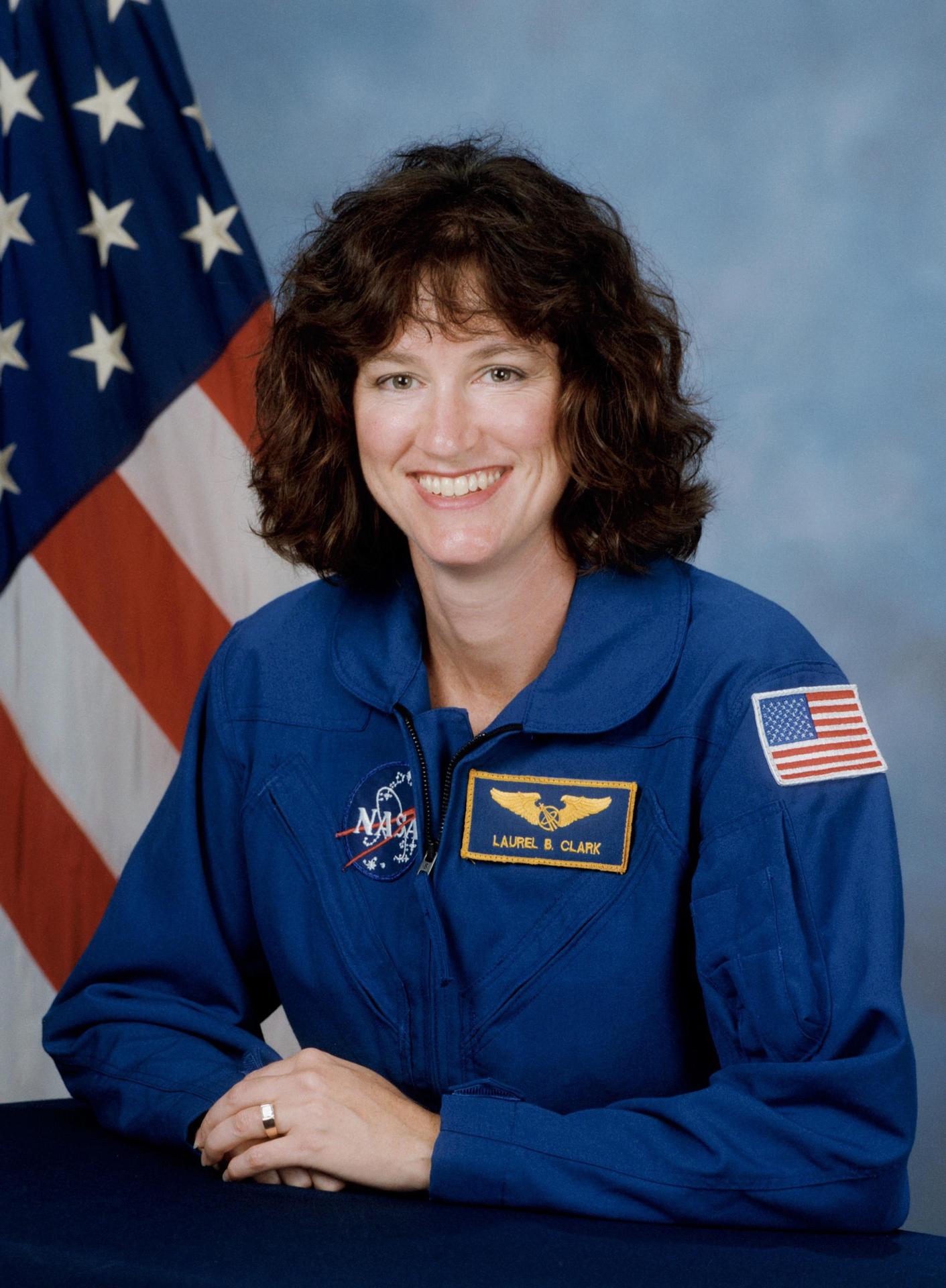 A woman with shoulder length hair smiles and sits in front of the American flag. She wears an astronaut flight suite.