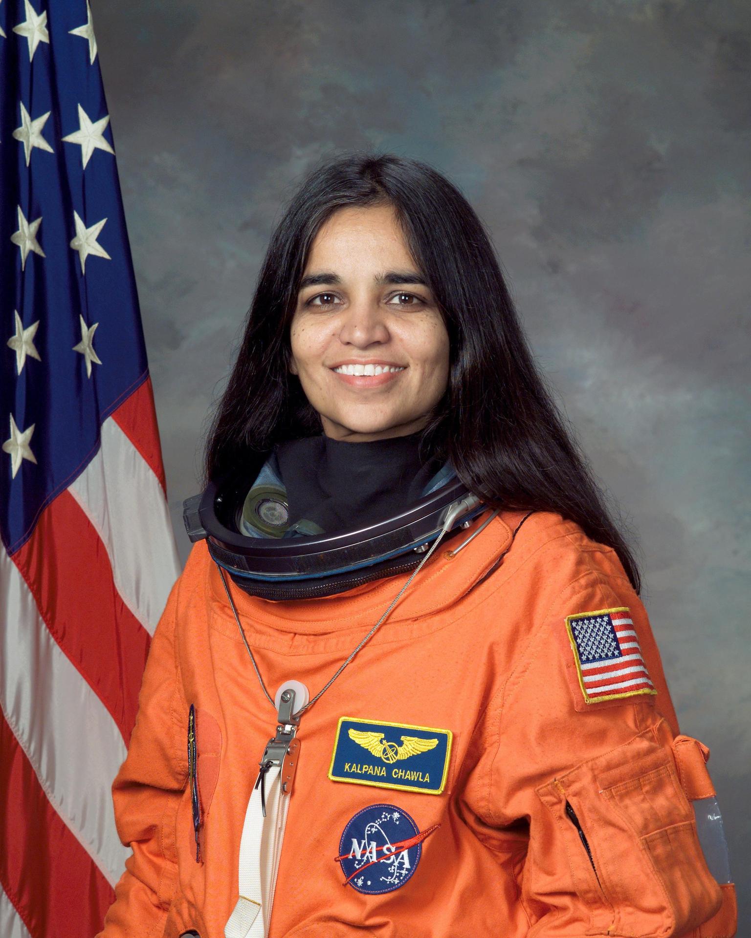 A woman with long straight hair, wearing an astronaut flight suite smiles and stands in front of the American flag.