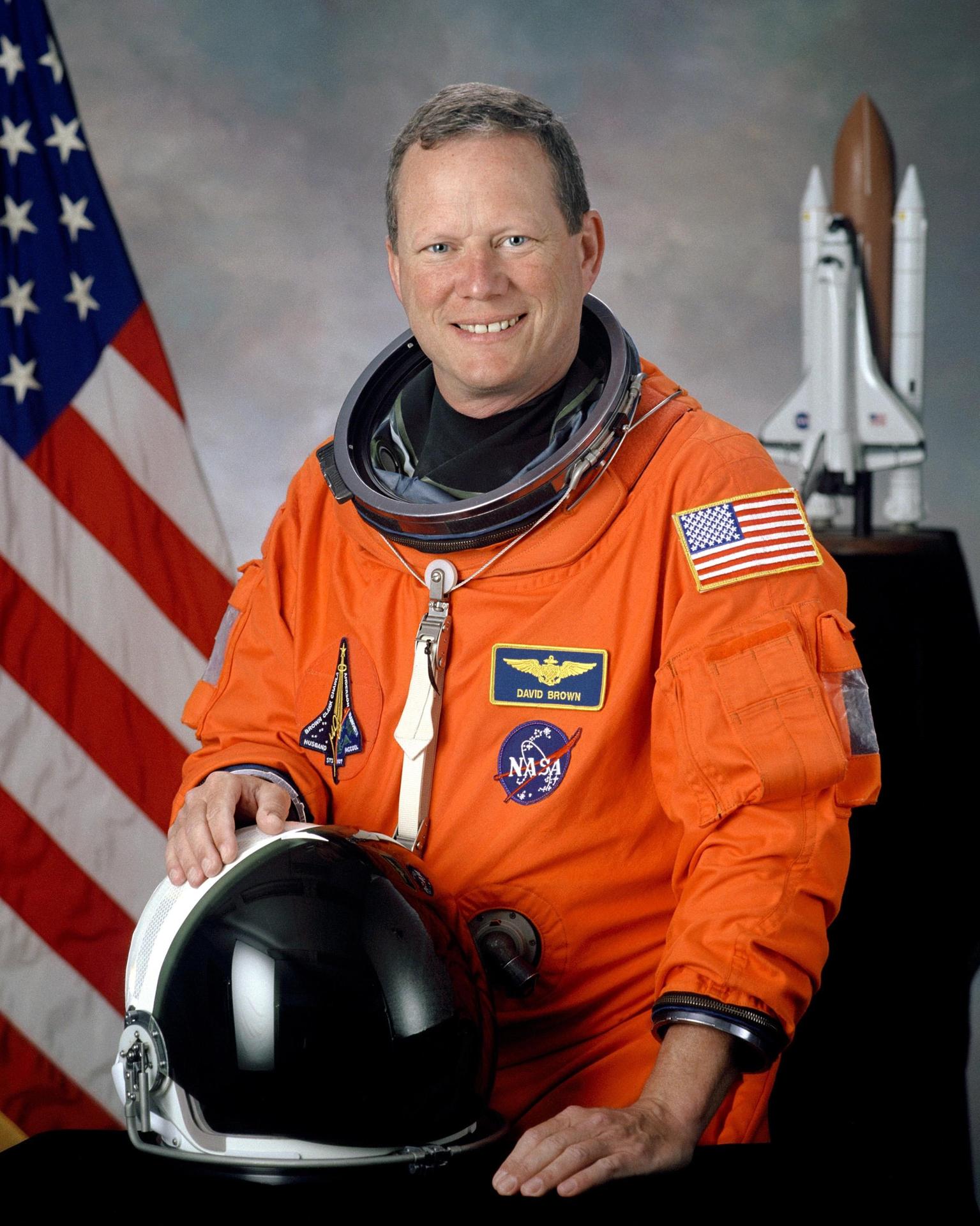 A man wearing a spacesuit smiles and stands in front of the American flag. He is holding his helmet in front of himself.