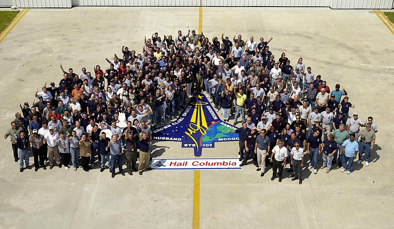 Members of the Columbia Reconstruction Project team pose for a group photo around an enlarged replica of the STS-107 crew emblem just delivered to the RLV Hangar. The emblem will be installed on an outside wall of the hangar. Inside the hangar, the team is identifying pieces of Columbia debris as they arrive at Kennedy Space Center and placing them on a grid approximating the shape of the orbiter.