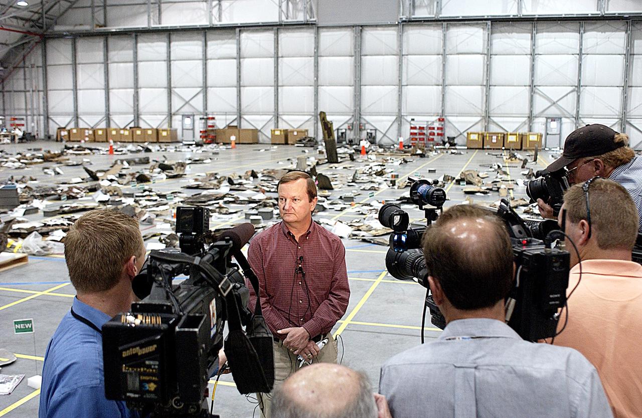 KENNEDY SPACE CENTER, FLA. -- Shuttle Launch Director Mike Leinbach answers reporters' questions in the RLV Hangar. Leinbach is overseeing assessment of the debris, seen in the background, as it is delivered to KSC. The Columbia Reconstruction Project team is identifying pieces and placing them on a grid approximating the shape of the orbiter. Shipped from Barksdale Air Force Base, Shreveport, La., more than 70,000 items, weighing 78,000 pounds, about 36 percent of the Shuttle by weight, have been delivered to KSC for use in the mishap investigation. Ground teams have completed 78 percent of their primary search area, and airborne crews finished 80 percent of their assigned area. Search teams have completed 98 percent of the underwater searches in Lake Nacogdoches and Toledo Bend Reservoir.