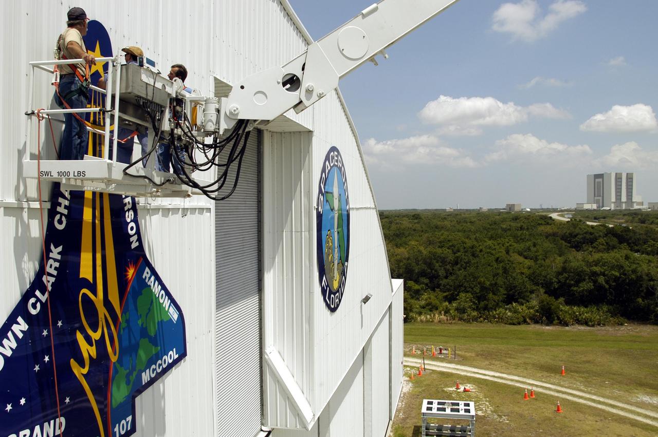 KENNEDY SPACE CENTER, FLA. - Workers place the final piece of the mission patch for STS-107 on the outside of the RLV Hangar at KSC.  The hangar is the site of the Columbia Reconstruction Project, where pieces of debris from Columbia are being collected and identified as part of the mishap investigation.  In the background, at right, is the Vehicle Assembly Building.