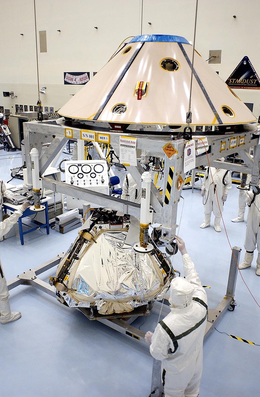 KENNEDY SPACE CENTER, FLA. - Workers in the Payload Hazardous Servicing Facility guide the backshell being lowered over the Mars Exploration Rover 1 (MER-1).  The backshell is a protective cover for the rover.  NASA's twin Mars Exploration Rovers are designed to study the history of water on Mars. These robotic geologists are equipped with a robotic arm, a drilling tool, three spectrometers, and four pairs of cameras that allow them to have a human-like, 3D view of the terrain. Each rover could travel as far as 100 meters in one day to act as Mars scientists' eyes and hands, exploring an environment where humans can't yet go.  MER-1 is scheduled to launch June 25 as MER-B aboard a Delta II rocket from Cape Canaveral Air Force Station.