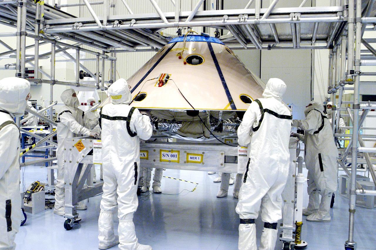 KENNEDY SPACE CENTER, FLA. -  Workers watch as an overhead crane begins to lift the backshell with the Mars Exploration Rover 1 (MER-1) inside.  The backshell will be moved and attached to the lower heat shield.  NASA's twin Mars Exploration Rovers are designed to study the history of water on Mars. These robotic geologists are equipped with a robotic arm, a drilling tool, three spectrometers, and four pairs of cameras that allow them to have a human-like, 3D view of the terrain. Each rover could travel as far as 100 meters in one day to act as Mars scientists' eyes and hands, exploring an environment where humans can't yet go.  MER-1 is scheduled to launch June 25 as MER-B aboard a Delta II rocket from Cape Canaveral Air Force Station.
