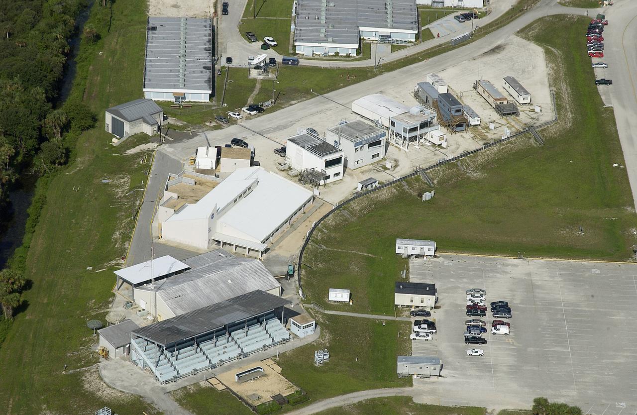 The KSC Press Site is seen from the air. The large white building is the NASA News Center. At lower left are the grandstands that provide viewing of Shuttle launches for the media. Stretching from the News Center to the right are buildings and trailers occupied by television media and other news agencies such as Associated Press and Reuters.