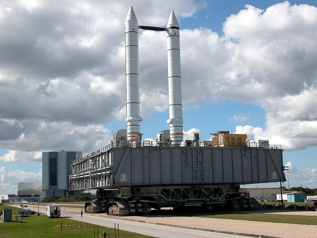 NASA image: KENNEDY SPACE CENTER, FLA. - A crawler-transporter carrying Mobile Launcher Platform (MLP) number 3, with a set of twin solid rocket boosters bolted atop, crawls to the intersection in the crawlerway in support of the second engineering analysis vibration test on the crawler and MLP.  From this perspective, the Launch Control Center (left) and the 525-foot-tall Vehicle Assembly Building (right) in the background appear dwarfed by the 184-foot-tall boosters. The crawler is moving at various speeds up to 1 mph in an effort to achieve vibration data gathering goals as it leaves the VAB, travels toward Launch Pad 39A and then returns. The boosters are braced at the top for stability. The primary purpose of these rollout tests is to gather data to develop future maintenance requirements on the transport equipment and the flight hardware. Various parts of the MLP and crawler transporter have been instrumented with vibration data collection equipment.