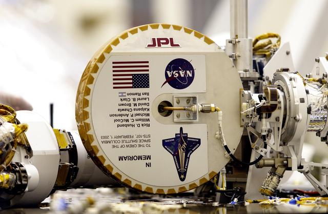 NASA image: KENNEDY SPACE CENTER, FLA. -- This plaque commemorating the STS-107 Space Shuttle Columbia crew now looks over the Mars landscape after the successful landing and deployment of the Mars Exploration Rover “Spirit” Jan. 4 onto the red planet.  The plaque, mounted on the high-gain antenna, is shown while the rover underwent final checkout March 28, 2003, in the Payload Hazardous Servicing Facility at KSC.