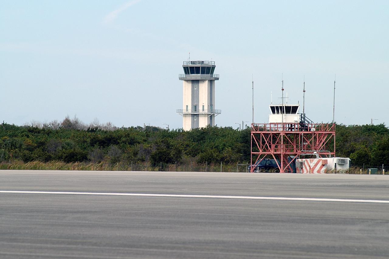 KENNEDY SPACE CENTER, FLA. -- Two control towers are seen at the edge of the KSC Shuttle Landing Facility, the old one in front and the nearly completed new tower in back.  The old tower stands only 20 feet above the runway surface, too low to see the launch pads to the east. During nighttime landing operations, those inside the tower have been hindered by the eight-billion candlepower xenon lights that illuminate the runway.  The new control tower is built atop an existing mound, rising nearly 100 feet over the midpoint of the runway.  The height gives controllers a spectacular 360-degree view of NASA-KSC and northern Brevard County. The new facility will also replace the SLF Operations Building. The operations building is home to the Military Radar Unit that monitors NASA-KSC airspace 24 hours a day, as well as runway light controls, navigational aids, weather and wind speed instrumentation, and gate controls. In the new tower, the computer displays will be fully modernized to Federal Aviation Administration standards with touch-screen technology. Construction on the new facility began in February 2003 and is nearly ready for occupancy. Only some final inspections and approvals remain. A support building and Public Affairs viewing deck, to be used for observing future landing operations, will be added and are already in work.