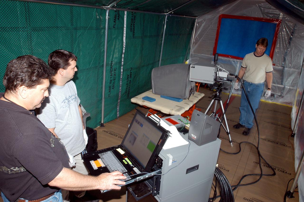 KENNEDY SPACE CENTER, FLA. -  Dan Phillips (left) and Donald Nielen, with United Space Alliance, watch a monitor as  Jim Landy, NDE specialist with USA, prepares to examine a Reinforced Carbon Carbon panel (on the table, center) using flash thermography.  A relatively new procedure at KSC, thermography uses high intensity light to heat areas of the panels. The panels are then immediately scanned with an infrared camera. As the panels cool, any internal flaws are revealed. The gray carbon composite RCC panels are attached to the leading edge of the wing of the orbiters.  They have sufficient strength to withstand the aerodynamic forces experienced during launch and reentry, which can reach as high as 800 pounds per square foot.  The operating range of RCC is from minus 250º F to about 3,000º F, the temperature produced by friction with the atmosphere during reentry.  The panels will be installed on the orbiter Discovery, designated  for the first Return to Flight mission, STS-114.