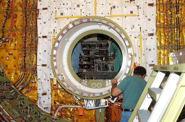NASA image: KENNEDY SPACE CENTER, FLA. -  A worker in the Orbiter Processing Facility checks the open hatch of the airlock in Discovery’s payload bay. The airlock is normally located inside the middeck of the spacecraft’s pressurized crew cabin. The airlock is sized to accommodate two fully suited flight crew members simultaneously. Support functions include airlock depressurization and repressurization, extravehicular activity equipment recharge, liquid-cooled garment water cooling, EVA equipment checkout, donning and communications. The outer hatch isolates the airlock from the unpressurized payload bay when closed and permits the EVA crew members to exit from the airlock to the payload bay when open.