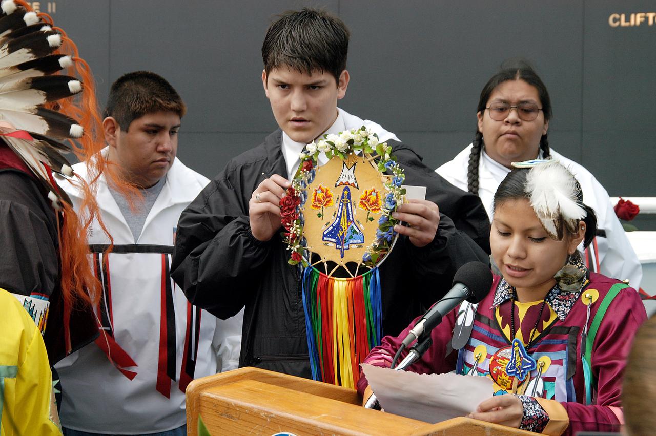 KENNEDY SPACE CENTER, FLA. - A member of the Shoshone-Bannock Native American community from Fort Hall, Idaho, reads a tribute to the crew of Columbia while another displays a handmade item with the STS-107 logo.  Dancers from Shoshone-Bannock Junior-Senior High School performed a healing ceremony during the memorial held at the Space Memorial Mirror, in the KSC Visitor Complex.  Feb. 1 is the one-year anniversary of the loss of the crew and orbiter Columbia in a tragic accident as the ship returned to Earth following mission STS-107.  Students and staff of the Shoshone-Bannock Nation had an experiment on board Columbia. The public was invited to the memorial service, held in the KSC Visitor Complex, which included comments by Center Director Jim Kennedy and Executive Director of Florida Space Authority Winston Scott.  Scott is a former astronaut who flew on Columbia in 1997.