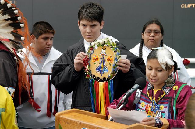 KENNEDY SPACE CENTER, FLA. - A member of the Shoshone-Bannock Native American community from Fort Hall, Idaho, reads a tribute to the crew of Columbia while another displays a handmade item with the STS-107 logo.  Dancers from Shoshone-Bannock Junior-Senior High School performed a healing ceremony during the memorial held at the Space Memorial Mirror, in the KSC Visitor Complex.  Feb. 1 is the one-year anniversary of the loss of the crew and orbiter Columbia in a tragic accident as the ship returned to Earth following mission STS-107.  Students and staff of the Shoshone-Bannock Nation had an experiment on board Columbia. The public was invited to the memorial service, held in the KSC Visitor Complex, which included comments by Center Director Jim Kennedy and Executive Director of Florida Space Authority Winston Scott.  Scott is a former astronaut who flew on Columbia in 1997.