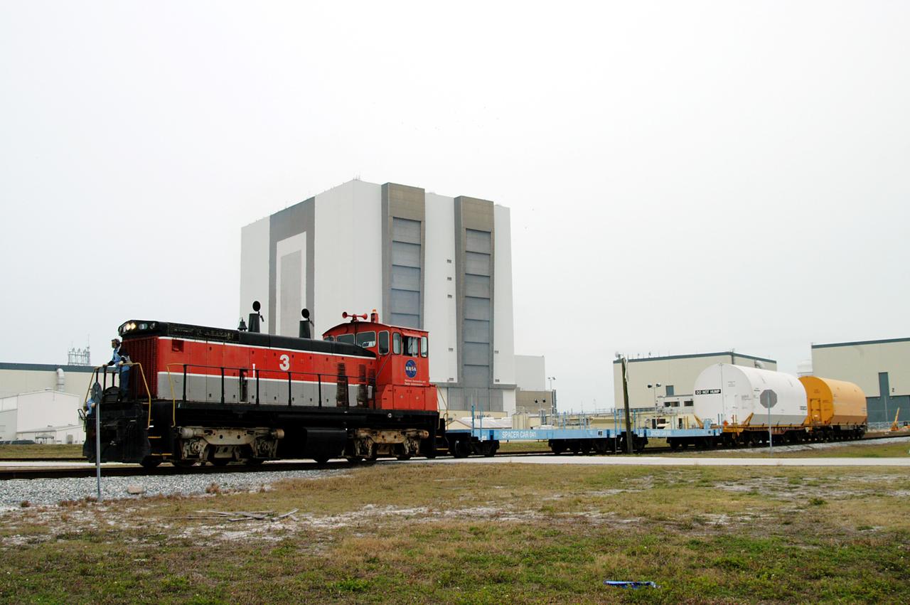 KENNEDY SPACE CENTER, FLA. - The red NASA engine moves forward past the Vehicle Assembly Building with its cargo of containers enclosing segments of a solid rocket booster being returned to Utah for testing.  The segments were part of the STS-114 stack.  It is the first time actual flight segments that had been stacked for flight in the VAB are being returned for testing.  They will undergo firing, which will enable inspectors to check the viability of the solid and verify the life expectancy for stacked segments.